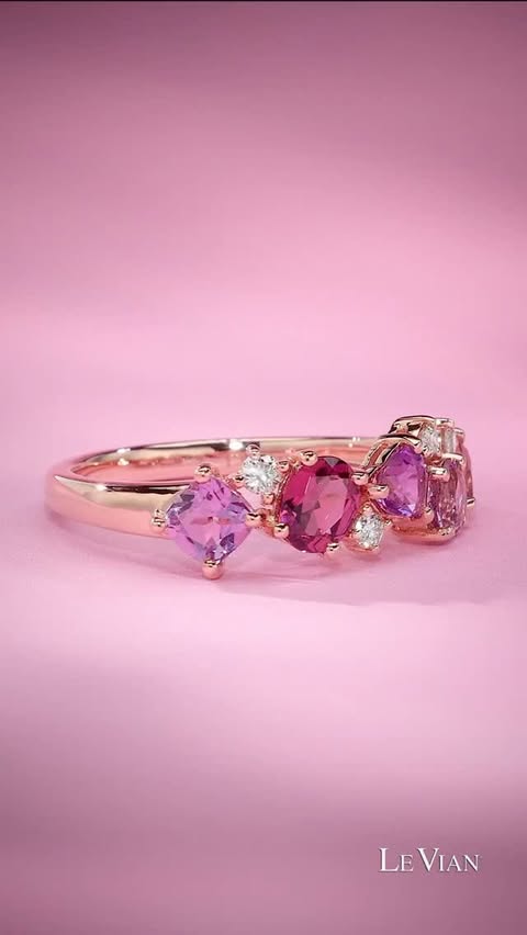 A ring rotates on a pink background. It has a cluster of gemstones in a line, from a heart-shaped amethyst to a pear-shaped garnet to passion fruit tourmaline to diamonds. It feels feminine and fun!