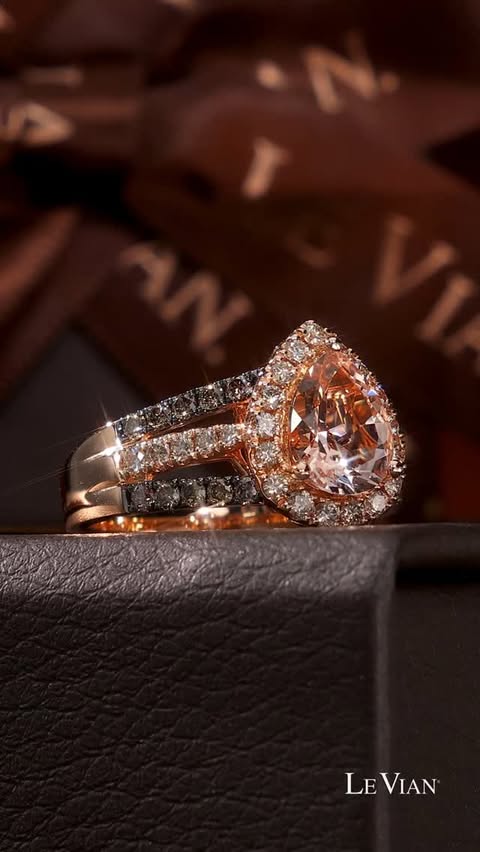 A peach morganite pear shaped gemstone in a ring with a triple row shank, adorned with diamonds in a halo and on the shank. It sits atop jewelry boxes.