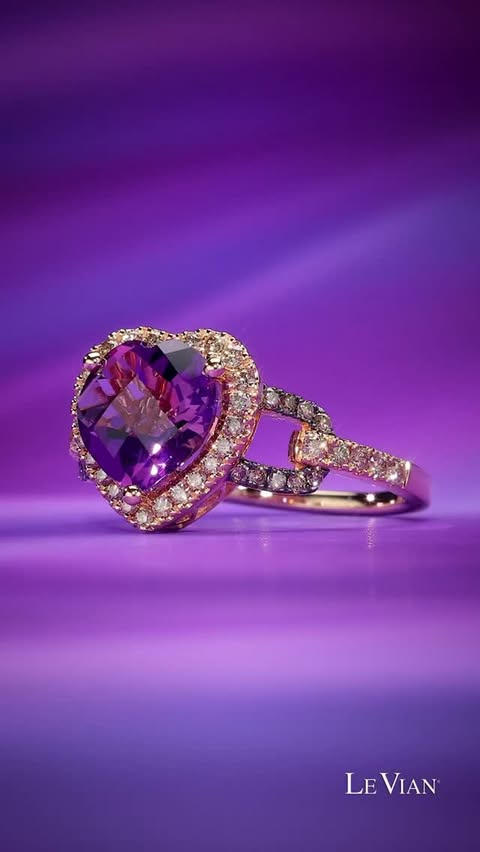 A purple grape amethyst center stone shaped like a heart sits within a diamond halo and a gold ring, turning with brilliance against a purple background.