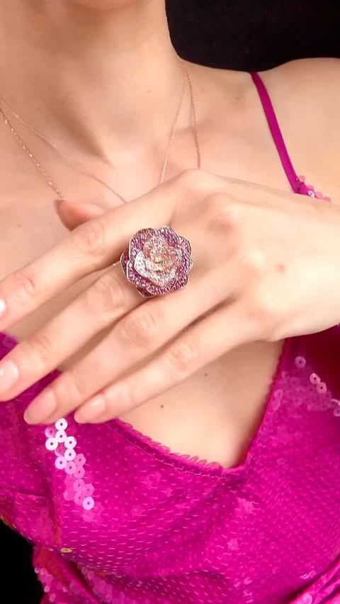 A woman wears ornate floral gemstone jewelry in pink ombré gemstones, in multiple necklaces and a ring. Her shirt is sparkly pink.